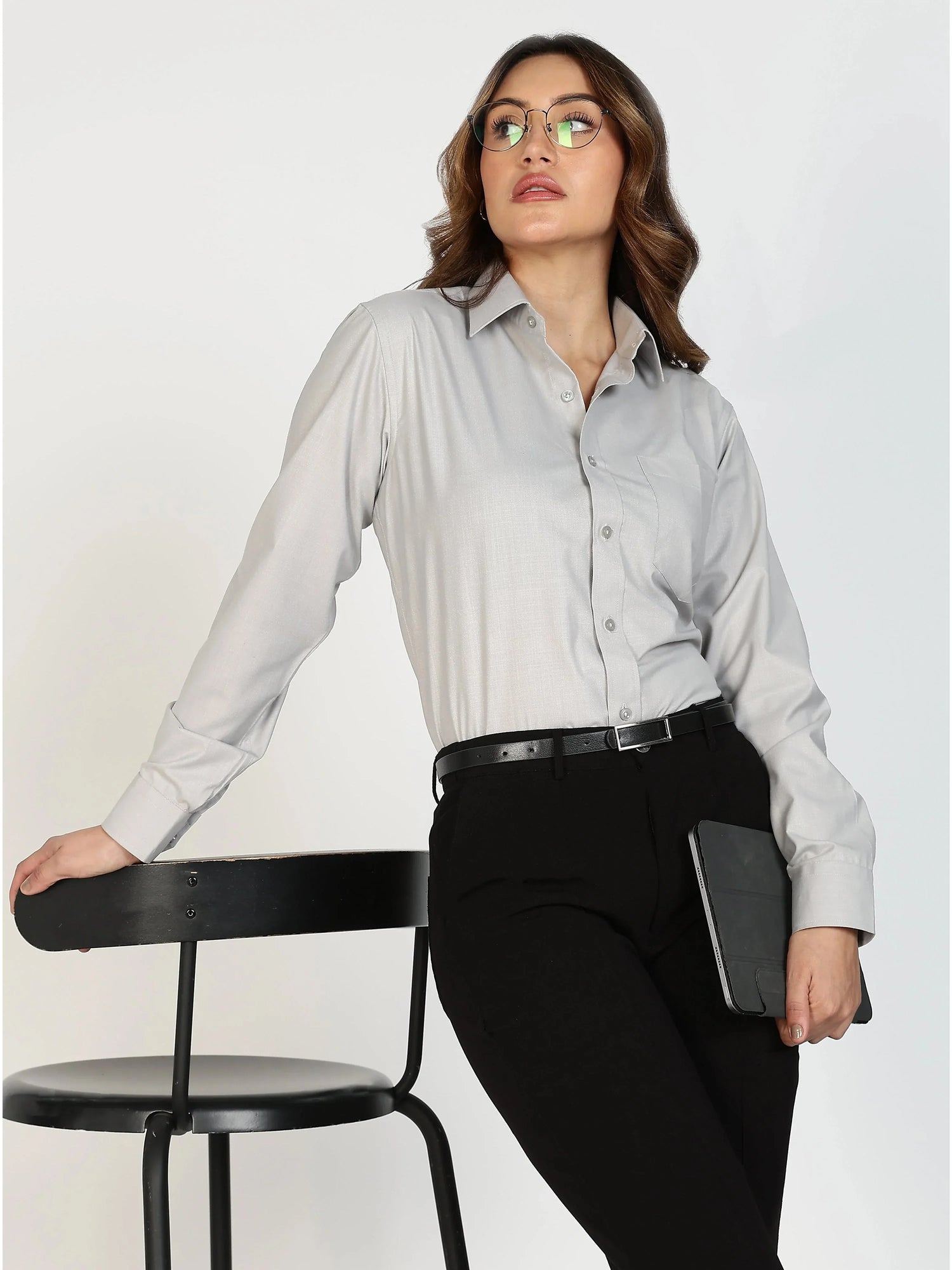 Woman in glasses wearing light gray formal shirt and black pants leaning on black chair holding tablet